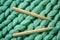 Two wooden knitting needles lie on a green chunky knitted merino wool