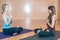 Two yound woman: a blond caucasian and an indian sitting in a lotus position in a yoga studio.