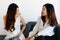 Two young Asian girls wearing office business style clothes sitting together on sofa, talking funny stories, smiling and laughing