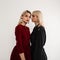 Two young attractive blonde women in fashionable clothes stand in studio near white vintage wall. Lovely girl in elegant black