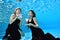 Two young beautiful girls, blonde and brunette, swim underwater in the pool with white cups in their hands, look at the camera and