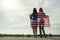 Two young friends women with USA national flag on their shoulders standing together outdoors on lake shore. Patriotic girls