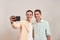 Two young happy caucasian twin brothers in casual wear smiling while taking selfie on mobile phone, standing together