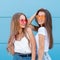 Two young hipster woman friends in retro neon sunglasses standing and smiling over blue wall