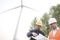 Two young male engineers in hardhats looking down at a blueprint in front of a wind turbine