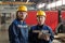 Two young serious engineers in protective hardhats and workwear looking at you in workshop