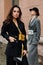 Two young stylish beautiful women fashion models are posing in street, wearing pantsuit, hat, having purse on waist.