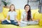 Two young teenagers sit in lotus pose on yellow sofa in room. They meditate. Girls express positive emotions. They keep