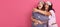 Two young woman in pajamas  on pink background with excited facial expressions, lady with curly hair pointing aside at