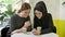 Two young womans sitting in cafe, one of them muslim woman in hijab, looking at phone and laughing, looking in camera