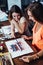 Two young women attending aquarelle painting classes for adults at art school. A girl showing her artwork to friend.