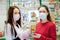 Two young women in medical masks and gloves on the background of a window with pharmacy products, with medicine in hand. The