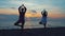 Two young women practice yoga by the sea or ocean at sunset.