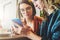 Two young women sit in a cafe at the table and use a smartphone. The girl shows her friend a picture on the phone screen