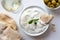 Tzatziki in white ceramic bowl with mint leaf garnish and a piece of pita bread next to pita bread, olives and a glass of white w