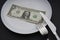 U.S. dollar money on a white plate, next to a knife and fork. Breakfast, lunch and dinner service.