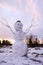 An ugly snowman with raised arms-branches stands in a snowy meadow at sunset