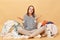 Uncertain confused Caucasian woman posing near heap of multicolored unsorted clothes isolated over beige background needs
