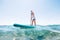 Under the water view angle to the smiling blonde teenager boy rowing stand up paddle board. Active family summer vacation time