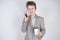 Unhappy angry teen in grey business suit stands with mobile phone and paper Cup of coffee on white background in Studio