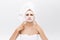 Unhappy beautiful young woman with white clay face mask makes acne therapy, wears white towel on hair, posing against
