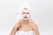Unhappy beautiful young woman with white clay face mask makes acne therapy, wears white towel on hair, posing against