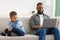 Unhappy Black Kid Sitting Near Reluctant Father Using Laptop Indoor
