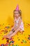 Unhappy blonde caucasian girl with boring face with scattered confetti. Yellow studio background. Bad birthday party