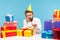 Unhappy bored man in cone hat holding cake with candle, sitting at workplace with many decorated gift boxes, lonely celebrating