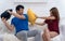 Unhappy couples battle of pillows on white couch living room. Angry couple fighting with color pillows at home