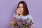 Unhappy dark haired woman suffers from runny nose uses nasal spray, has allergic rhinitis reaction on lilac flowers, posing