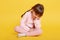 Unhappy little Caucasian girl sitting on floor isolated over yellow background, child with crossed legs feels boring, sits with