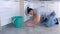 Unhappy tired man in rubber gloves washes and rubs hard the floor in the kitchen, sitting on the floor, side view.