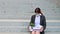 Unhappy woman in a mask sits with a box of personal stuff on the stairs outdoors. A female office employee was fired