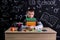 Unhappy youngster sitting at the desk with a pile of books under the chin, surrounded with school supplies. Chalkboard