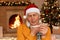 Unhealthy senior man in santa hat sitting near fireplace wrapped in scarf, holding thermometer in hands, has fever, posing in