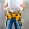 unidentified handyman standing with a tool belt with construction tools and holding project plan against grey background. DIY too