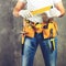 unidentified handyman standing with a tool belt with construction tools and holding roulette against grey background, toned image