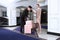 Uniformed doorman meets guest at hotel and puts her suitcases on luggage cart. Fashionable modern girl in pink business