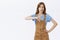 Unimpressed young pretty woman with chestnut hairstyle in overalls holding hand on hip showing thumb down in dislike