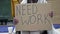 An unkempt-looking young woman sits with a begging cup and holds a handwritten NEED WORK poster.