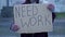 An unkempt-looking young woman sits with a begging cup and holds a handwritten NEED WORK poster.