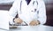 Unknown male doctor sitting and working with clipboard of medication history record in clinic at his working place