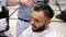 Unparalleled barber with a beard and a tattoo is cutting the hair of his client in the barbershop.