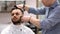 Unparalleled barber with a beard and a tattoo is cutting the hair of his client in the barbershop.