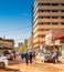 Unpaved side street with a skyscraper and small sales huts and stalls as well as cars and pedestrians in the middle of the street