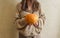An unrecognizable brunette girl in a beige sweater holds a bright orange pumpkin in her hands on a beige background.