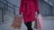 Unrecognizable elegant lady in red coat, gloves, and black boots walking with shopping bags in city. Stylish young woman