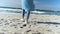 Unrecognizable female in sky blue coat, jeans and shoes walk alone on beach with white sand at autumn sunny day.