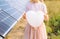 Unrecognizable girl child with white empty heart shaped balloon, standing next to solar panels, solar farm on sunny summer day.